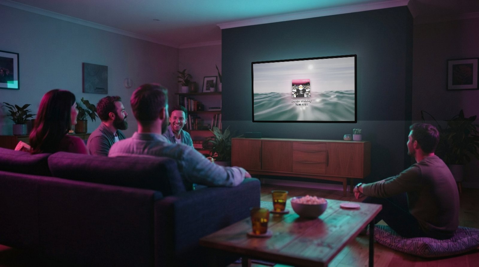 A living room at night, five friends on the sofa; on the wall-mounted TV, Halofloor visuals with the cover of Dance Monkey by Tones And I, its light spilling onto the wall.