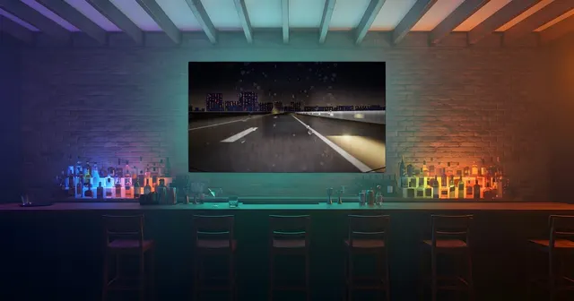 Bar de noche: pared de ladrillo, estanterías de botellas iluminadas de colores, taburetes alineados ante la barra y una gran pantalla encima con una carretera nocturna.