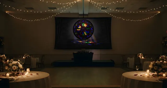 Salón de banquetes de boda por la noche: mesas redondas con mantel, centros de flores y velas, guirnaldas de luces en el techo, una pista de baile y una gran pantalla de proyección al fondo.